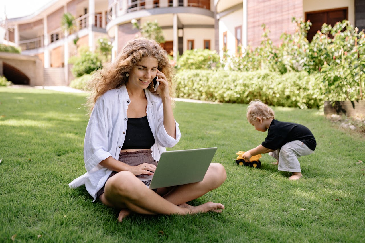 about-img Woman multitasking in garden with laptop and phone, child playing nearby.