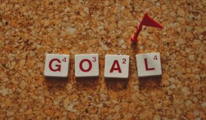 Scrabble tiles forming the word 'goal' with a small red flag on a cork board background.
